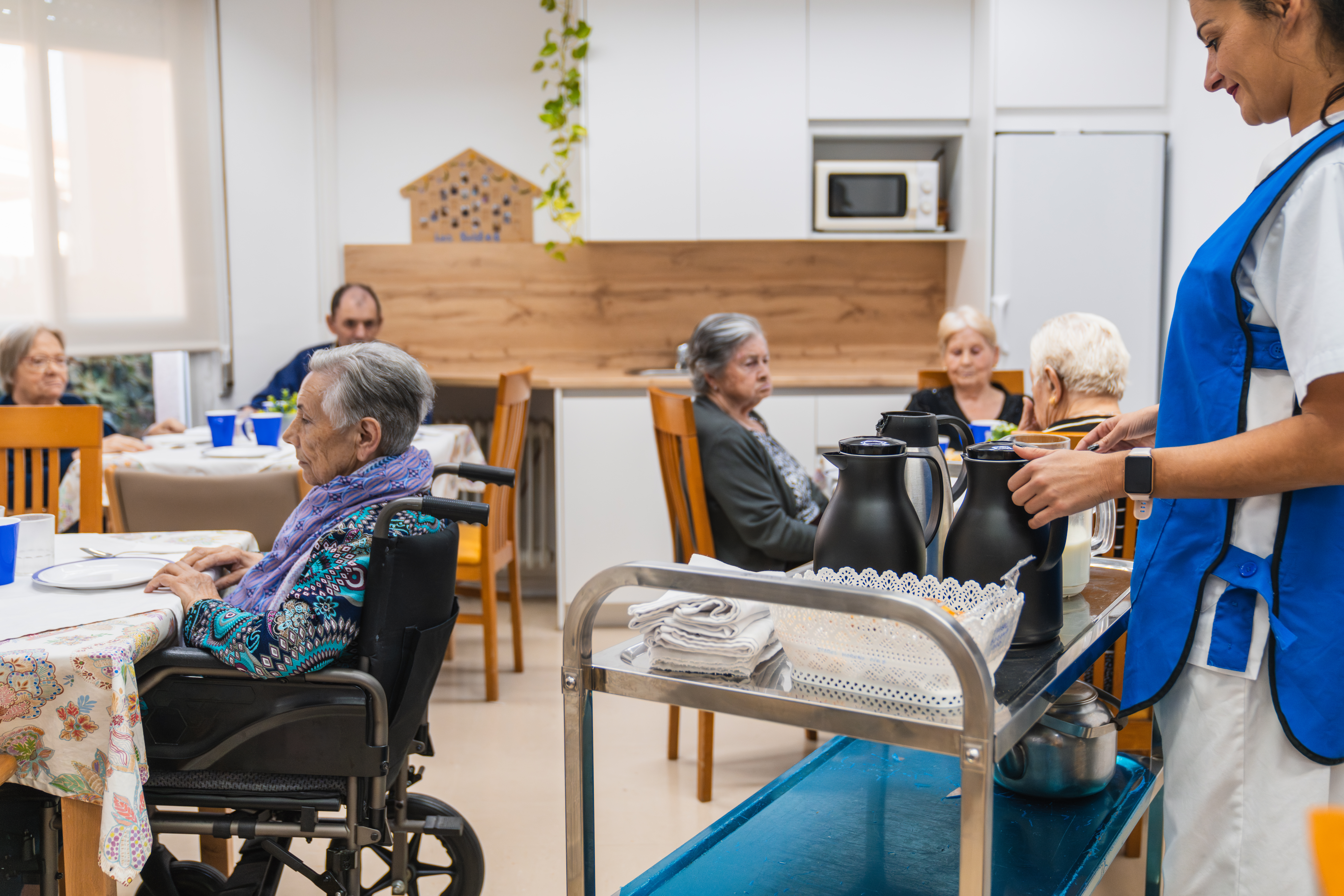 Nurse pushing cart serving elderly people in nursing home dining room