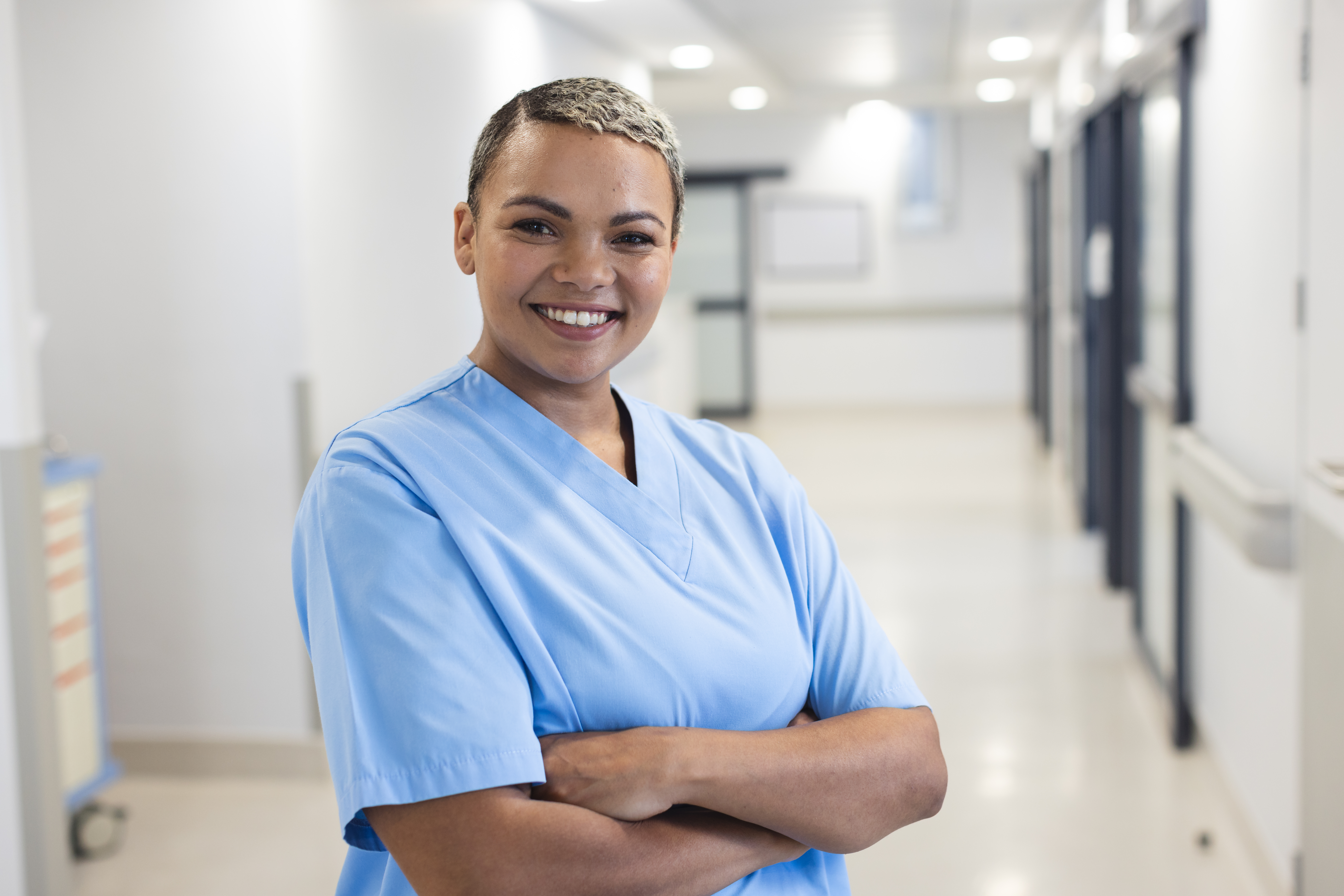 Portrait of happy biracial female doctor with short hair wearing scrubs in corridor at hospital. Medicine, healthcare, work and hospital, unaltered.