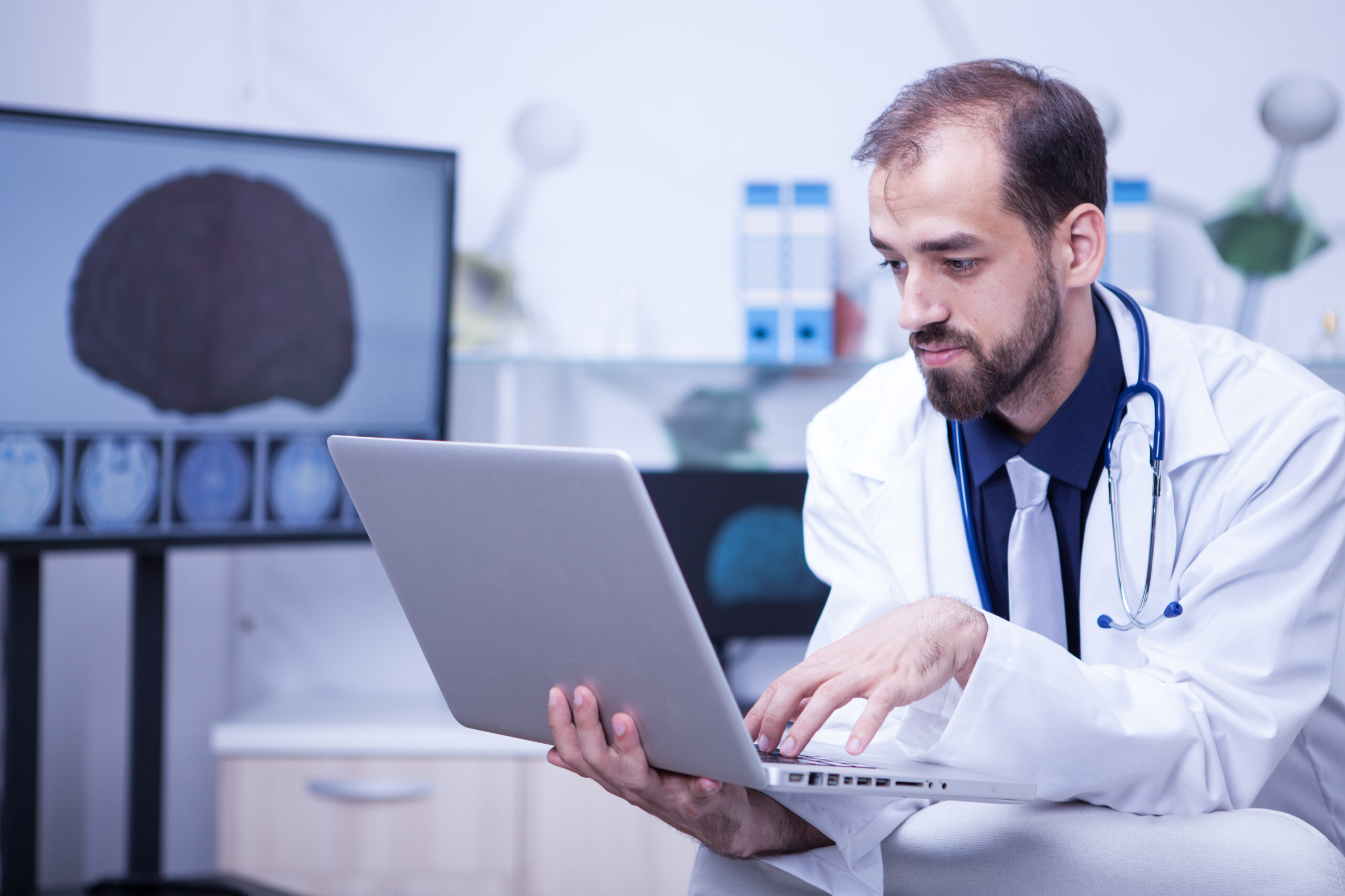 Attractive bearded doctor typing a diagnosticin in the patient file on his laptop
