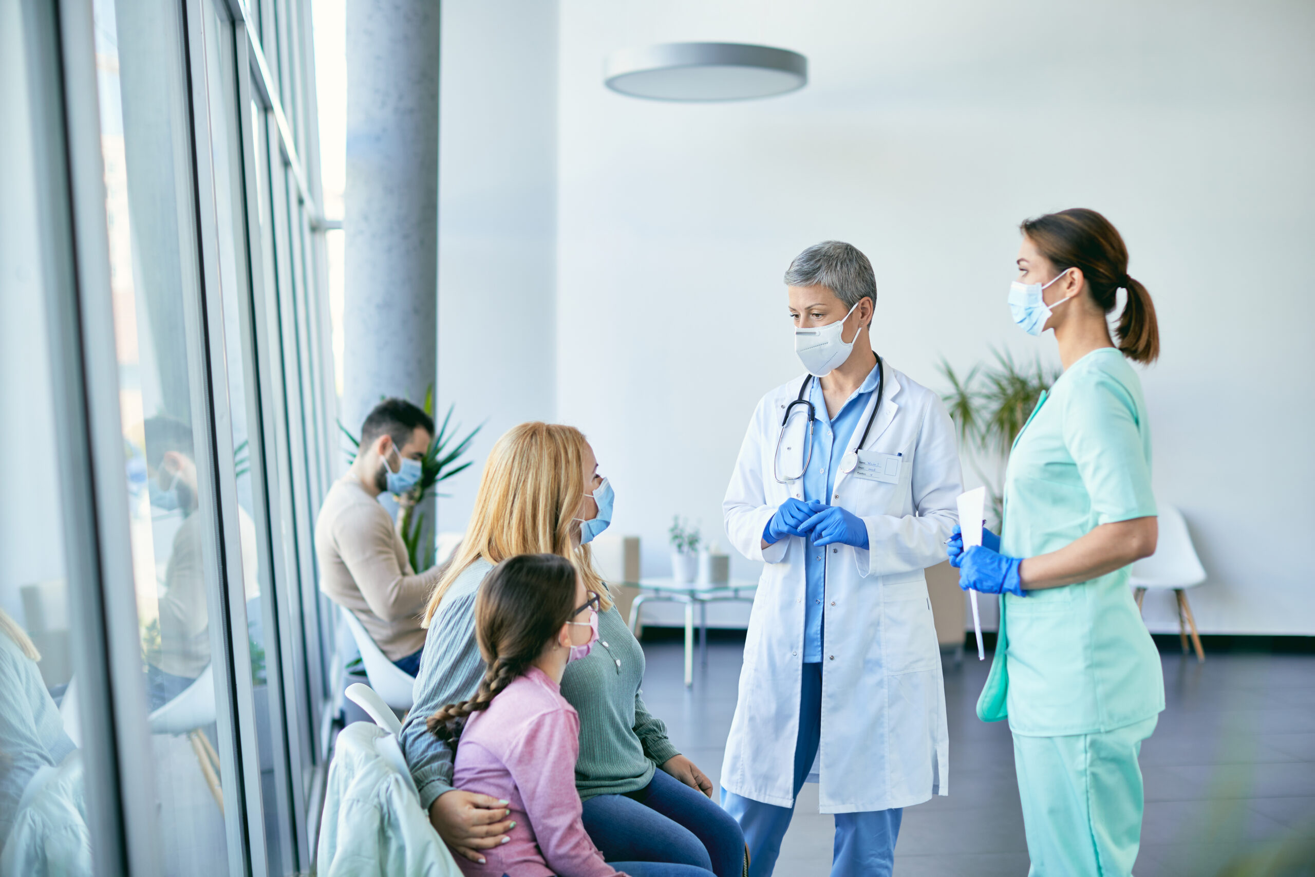 Mother and daughter communicating with doctor and nurse in waiting room at medical clinic during coronavirus pandemic.