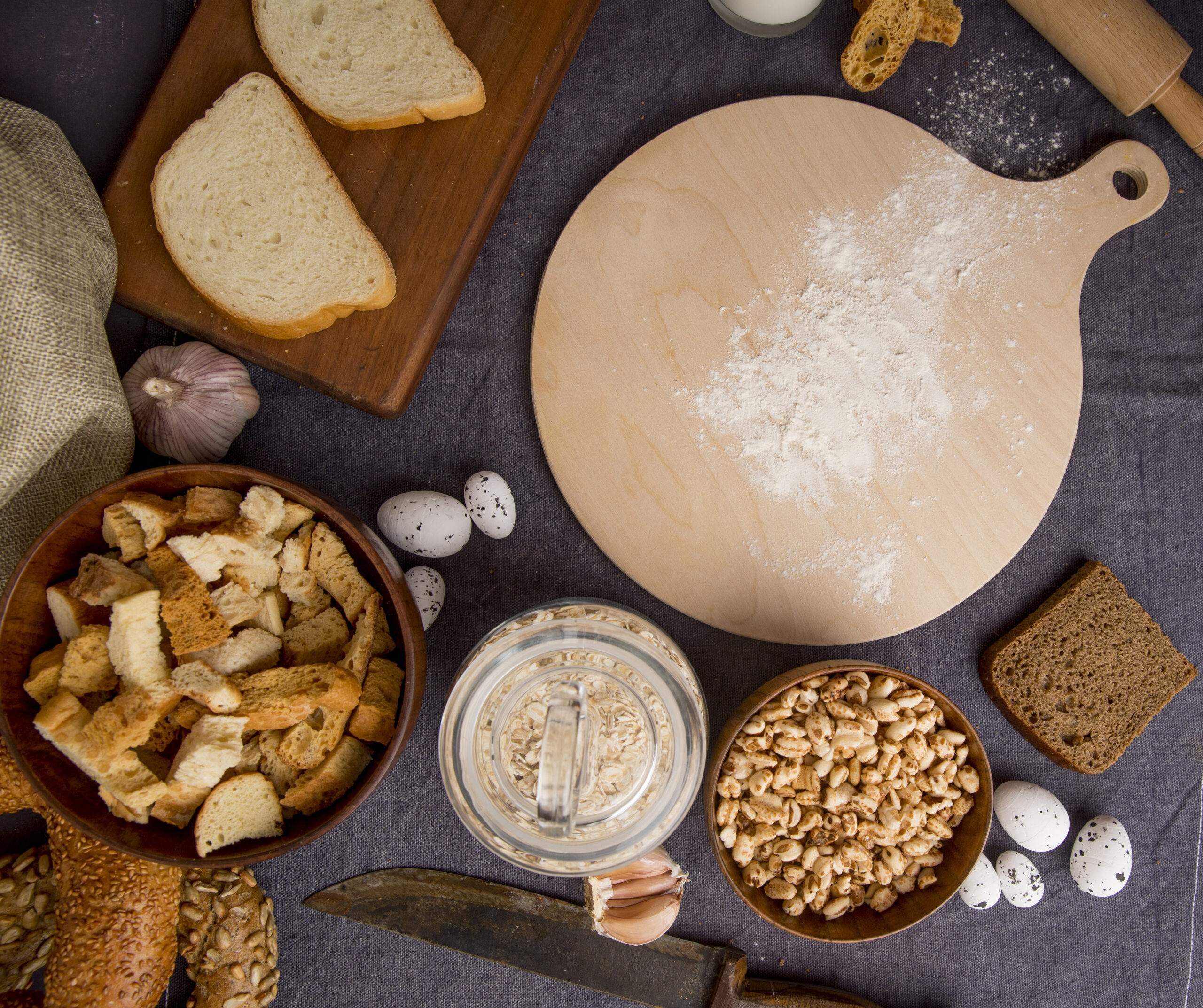 top view of bread pieces, oat-flakes, corns, garlic, eggs, knife with cutting board on maroon background