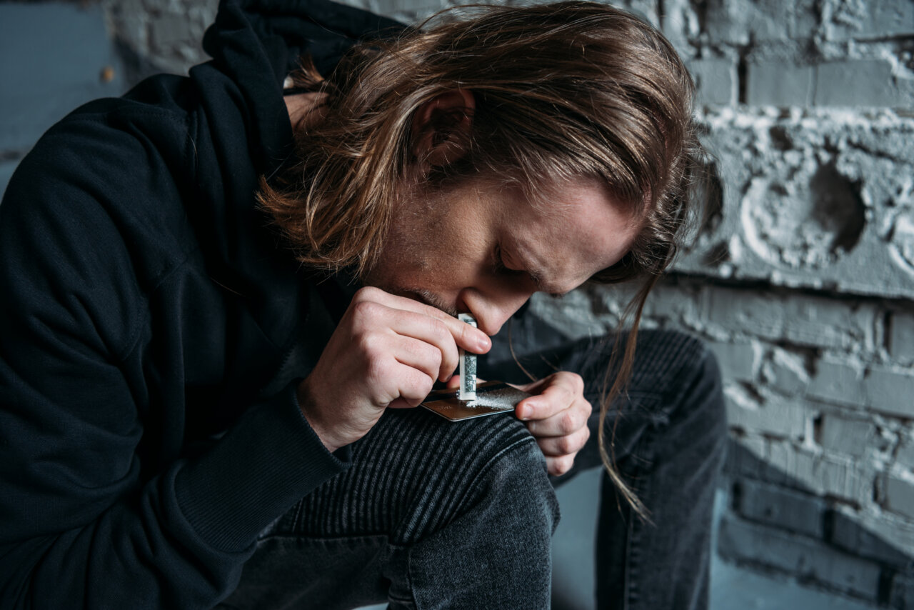 Hombre con cabello largo consumiendo cocaína en un entorno oscuro.